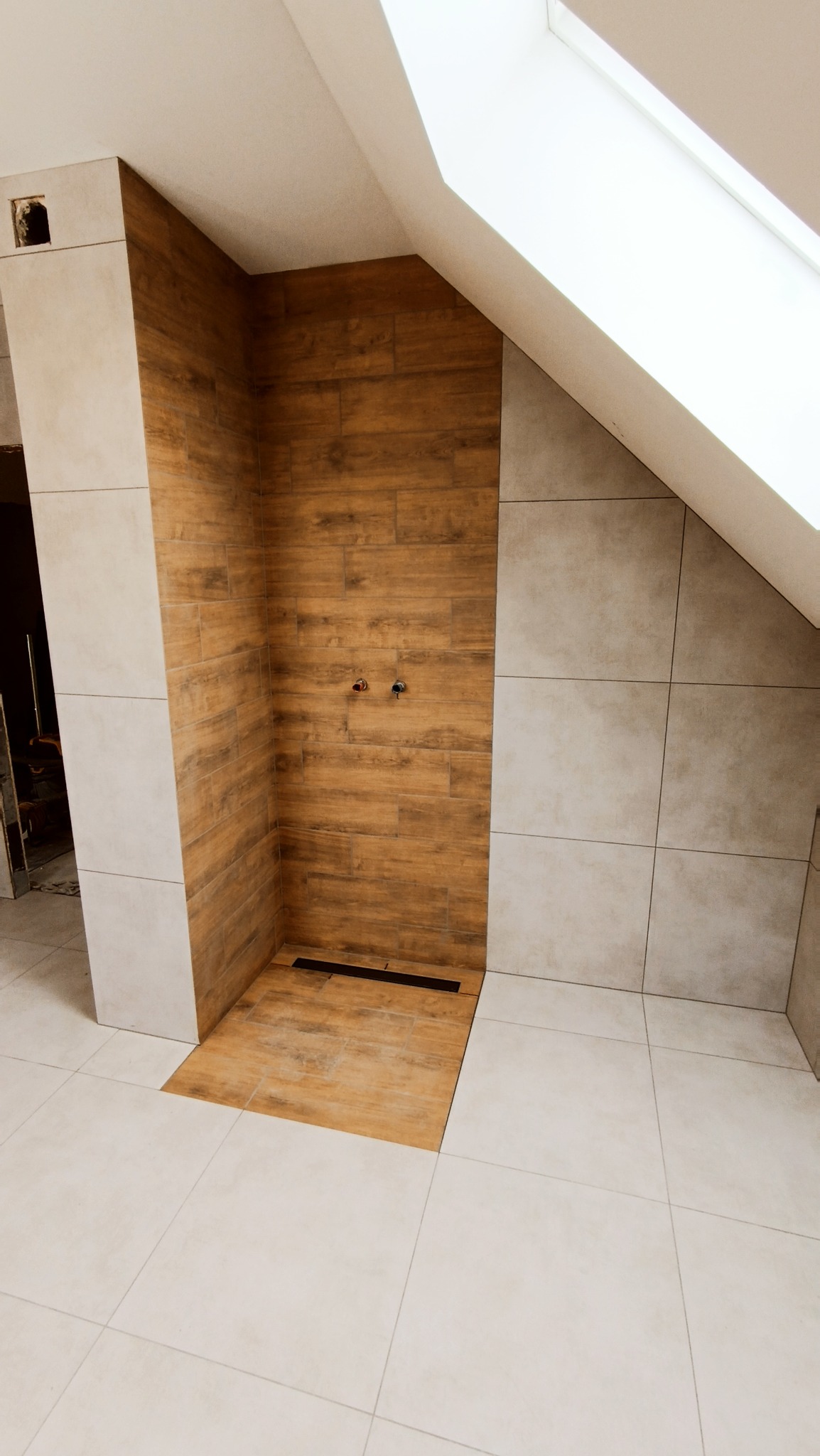 Niedokończona kabina prysznicowa z drewnopodobnymi płytkami, widoczne przyłącza wodne i odpływ liniowy, jasne płytki na podłodze i ścianach, okno dachowe.