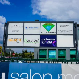 Billboard reklamowy z logo różnych firm, w tym agencji interaktywnej 'Ciekawa Strona', na tle budynku i błękitnego nieba z chmurami.