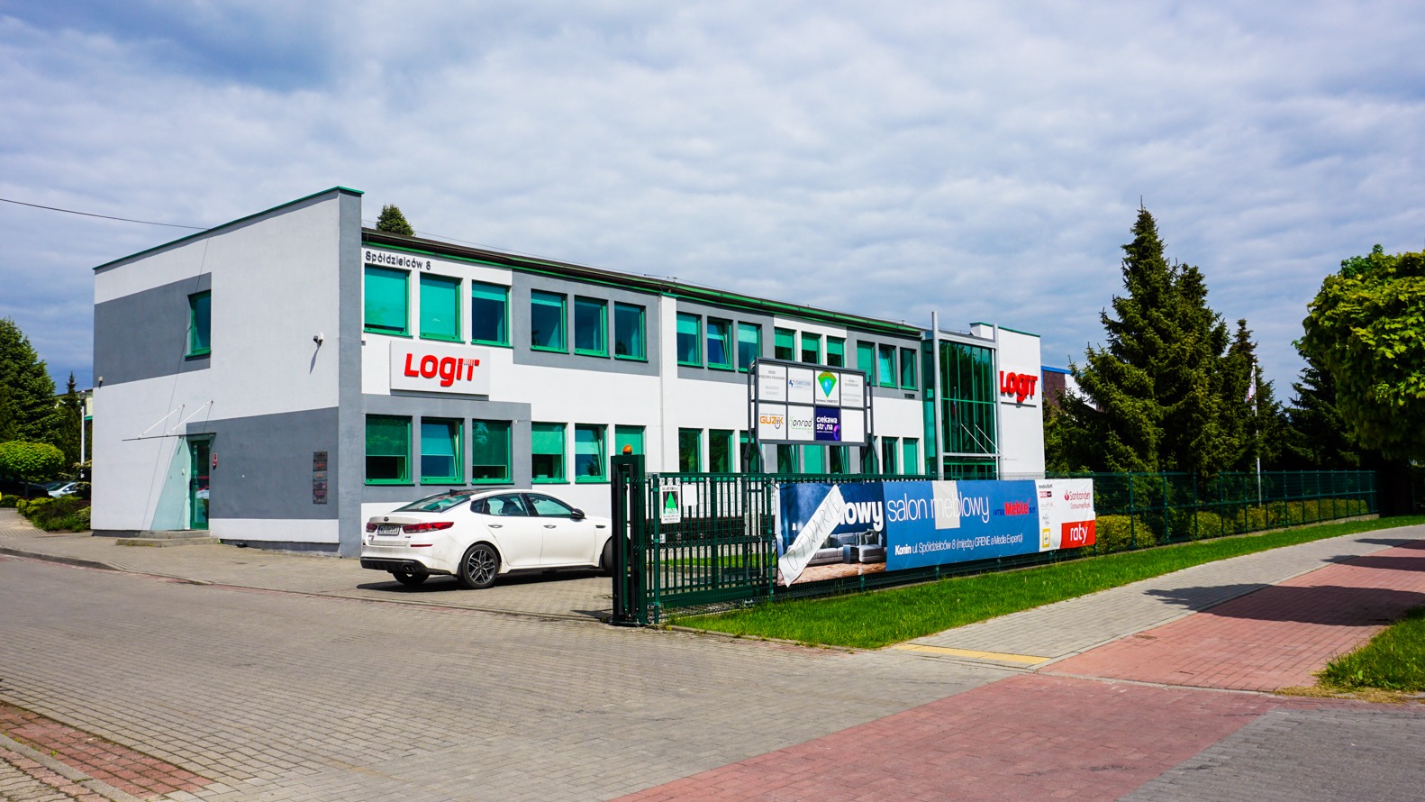 Budynek firmy Logit z czerwonym logo na białej fasadzie, widoczny baner reklamowy salonu meblowego i zaparkowany biały samochód.