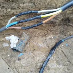 Wykop z ziemi z widocznymi kablami elektrycznymi zabezpieczonymi czarną taśmą izolacyjną oraz niebieską rurą karbowaną, fragment drewnianej deski i błoto.