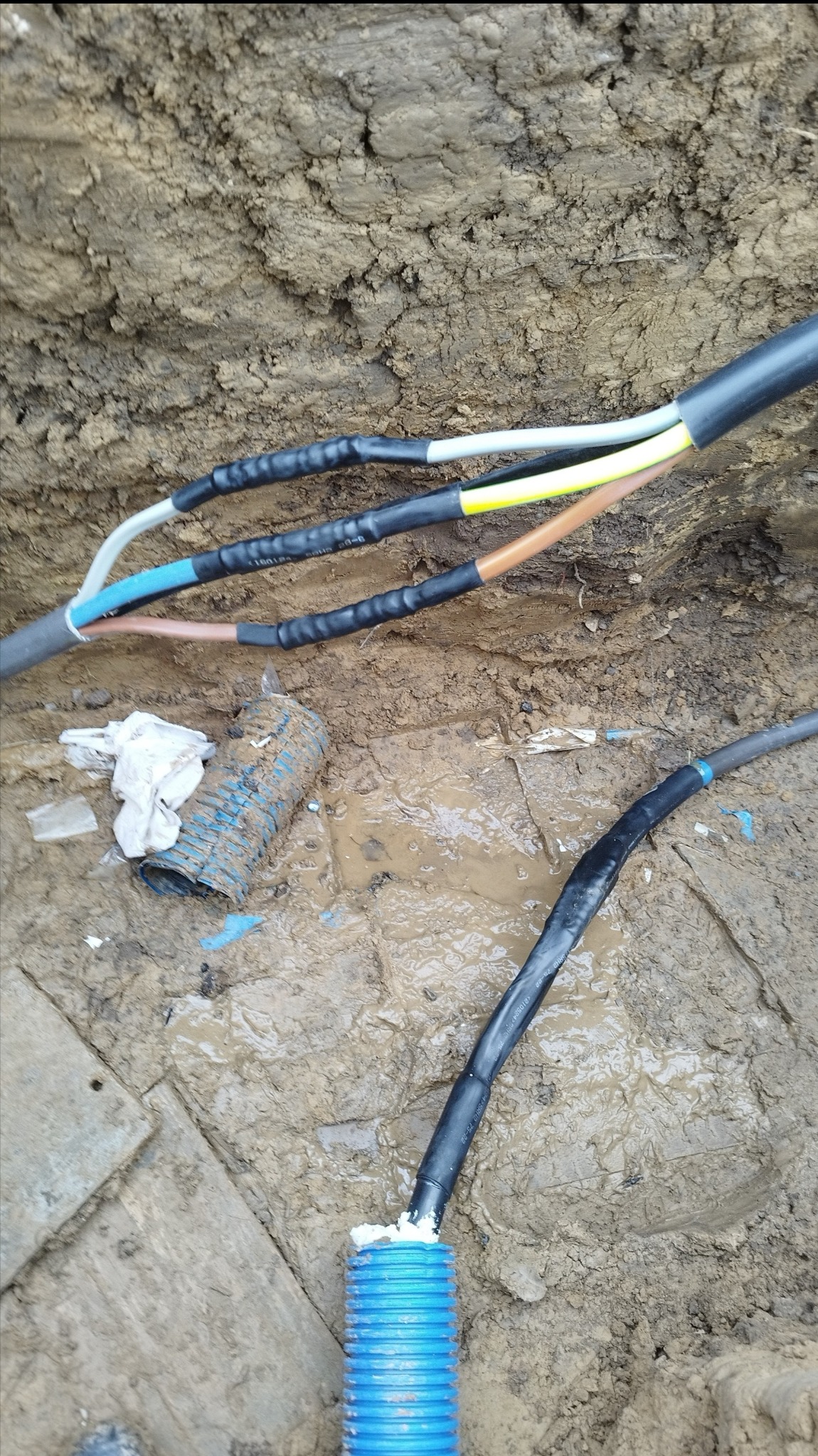 Wykop z ziemi z widocznymi kablami elektrycznymi zabezpieczonymi czarną taśmą izolacyjną oraz niebieską rurą karbowaną, fragment drewnianej deski i błoto.
