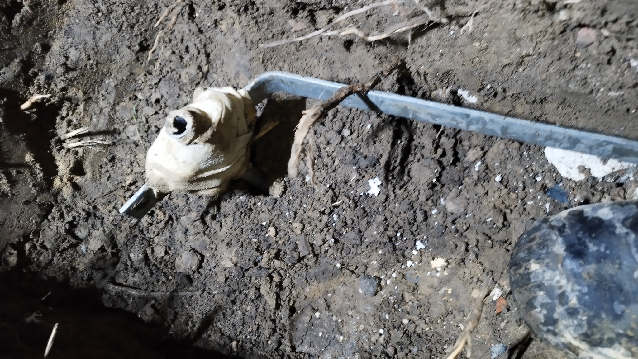 Podłączenie rury w wykopie ziemnym, owinięte materiałem izolacyjnym, z widocznymi korzeniami i elementami instalacji.