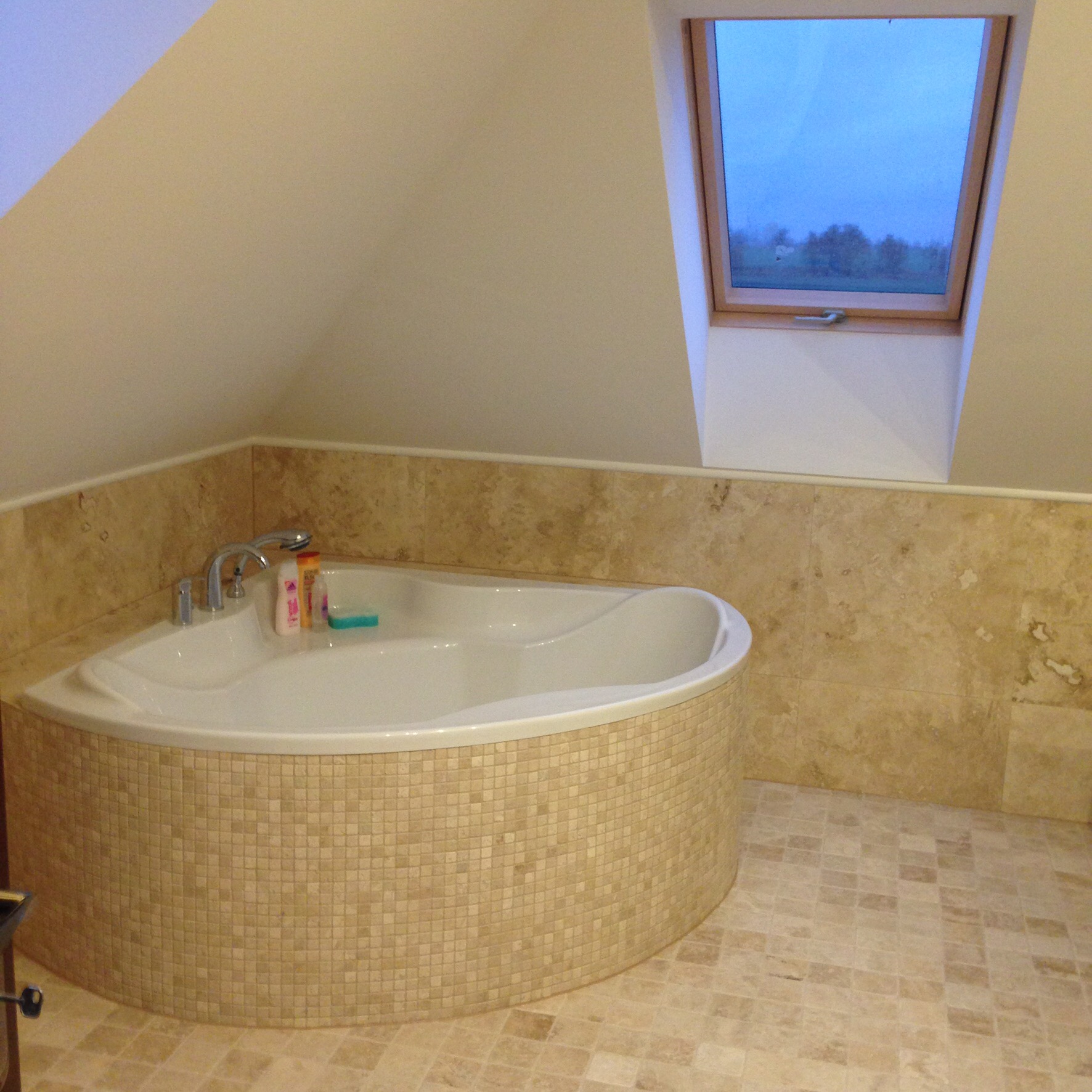 Narożna wanna obudowana mozaiką w beżowych odcieniach, z baterią i kosmetykami, na tle ściany wyłożonej płytkami imitującymi trawertyn oraz okna dachowego z widokiem na zewnątrz.