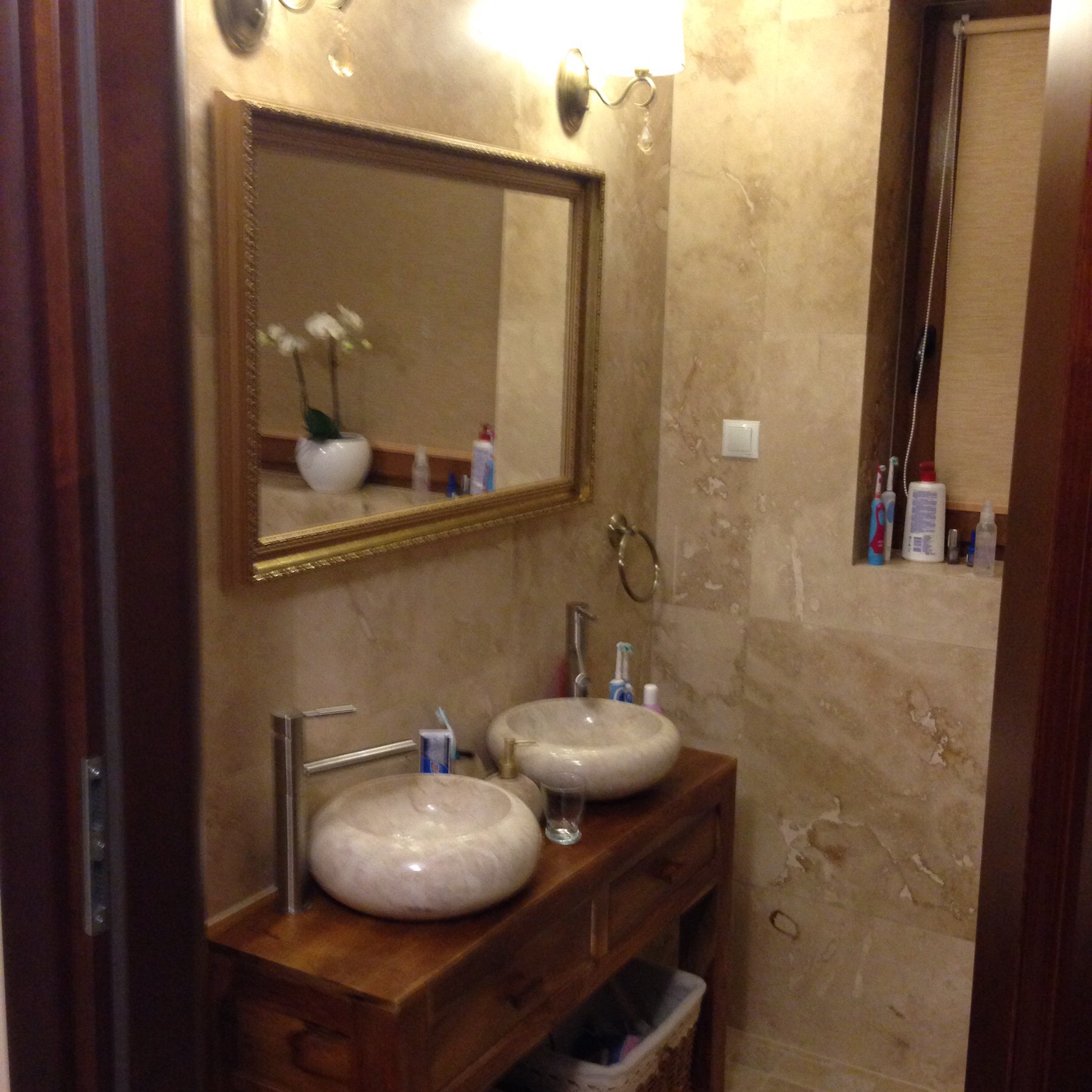 Wykończona łazienka z dwoma okrągłymi umywalkami z kamienia na drewnianej szafce, lustro w złotej ramie i beżowe płytki imitujące marmur.