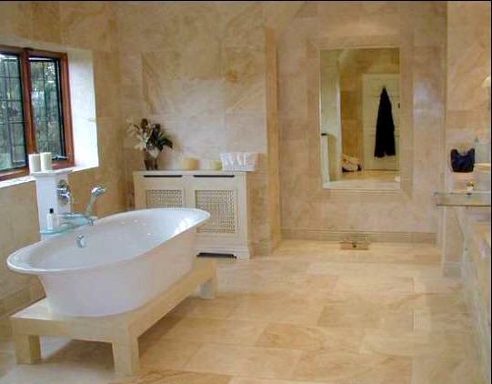 Elegancka łazienka z wanną wolnostojącą na podwyższeniu, wykończona marmurem w odcieniach beżu, z dużym lustrem i oknem z widokiem na zieleń.