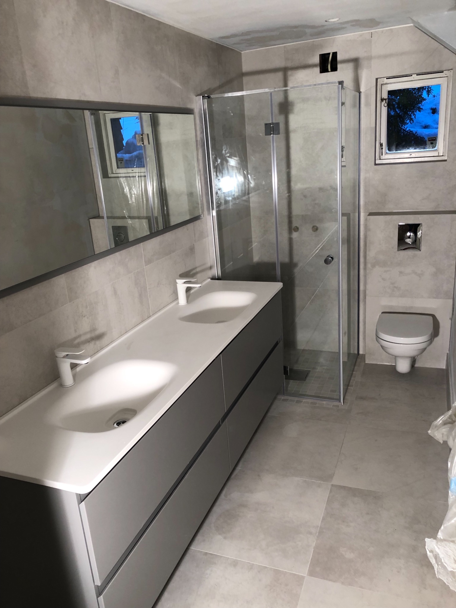 Szara łazienka po remoncie: podwójna umywalka z szafką, kabina prysznicowa, toaleta podwieszana i okno z widokiem na drzewa.