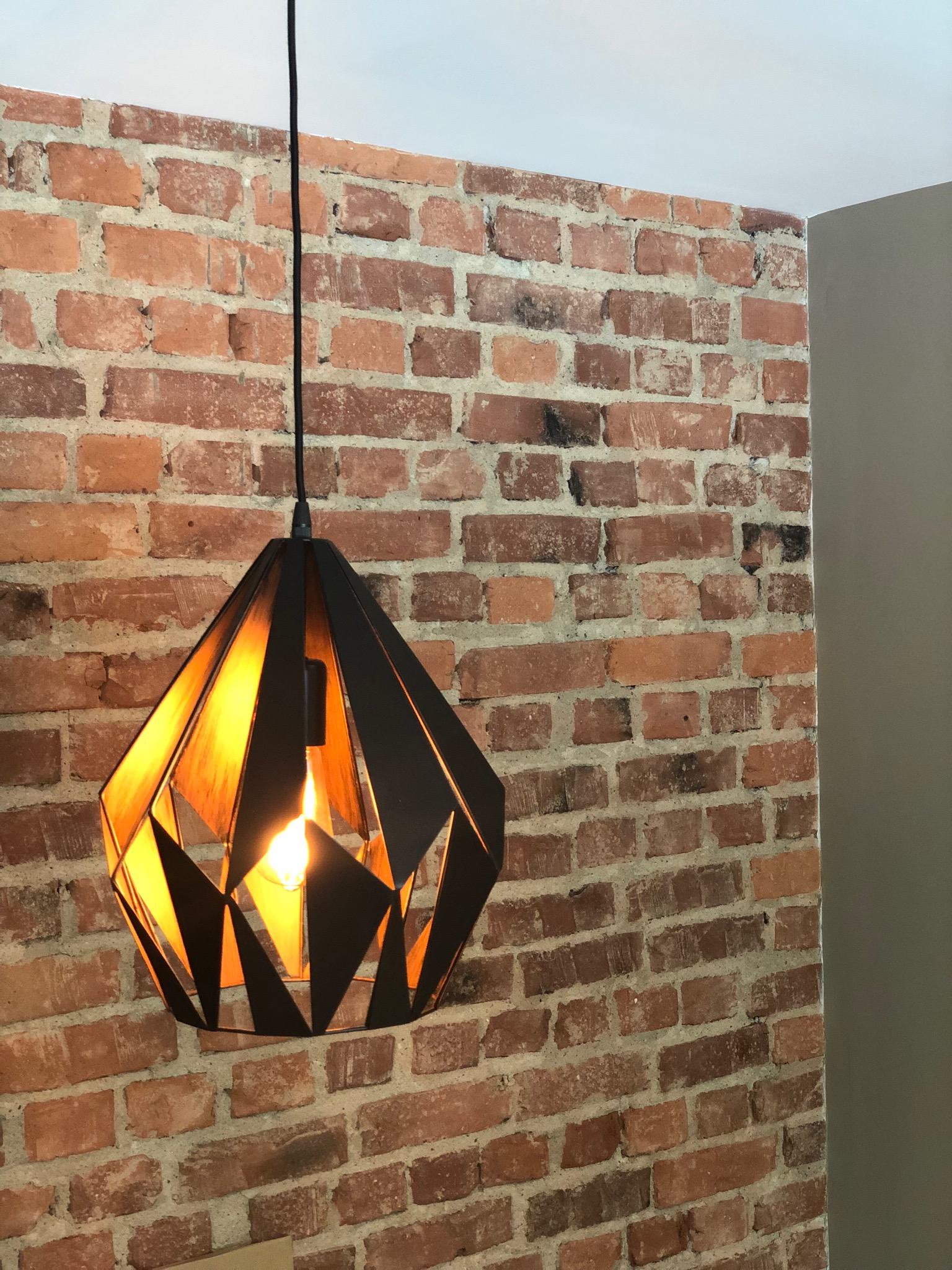 Lampa wisząca o geometrycznym, czarnym kloszu z widoczną żarówką na tle ściany z czerwonej cegły w pomieszczeniu.