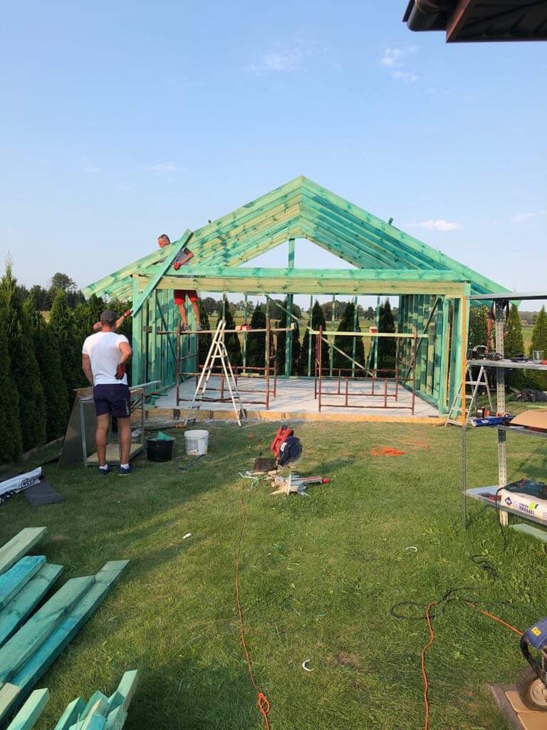 Budowa drewnianej altany ogrodowej w trakcie realizacji: widoczna konstrukcja szkieletowa z impregnowanego drewna, pracownicy na drabinie mocują elementy, na trawie leżą deski i narzędzia.