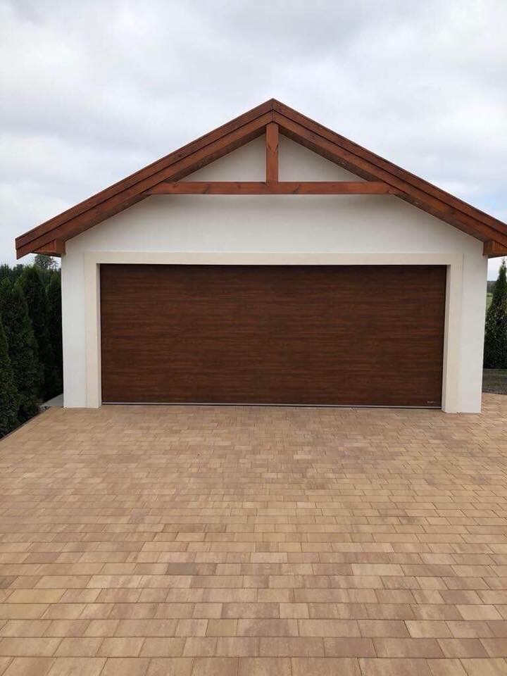 Nowoczesny garaż z brązową bramą segmentową i drewnianym zadaszeniem, wykończony jasną elewacją i brukowanym podjazdem.