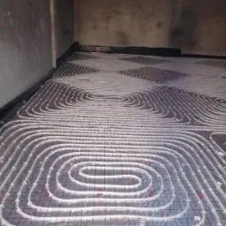 Montaż rur grzewczych podłogowych na folii z siatką w surowym wnętrzu, widoczne pętle i fragment ściany.