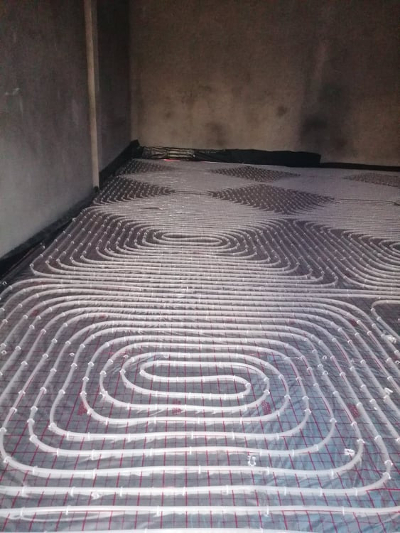 Montaż rur grzewczych podłogowych na folii z siatką w surowym wnętrzu, widoczne pętle i fragment ściany.