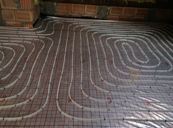 Rozłożone białe rurki ogrzewania podłogowego na folii z czerwoną siatką, widoczne elementy konstrukcji sufitu.