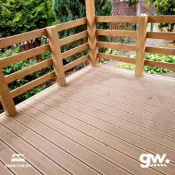 Kątowe ujęcie nowo wybudowanego tarasu z desek kompozytowych, z drewnianą balustradą w naturalnym odcieniu, otoczonego zielenią ogrodu. Widoczne logo producenta desek w rogu.