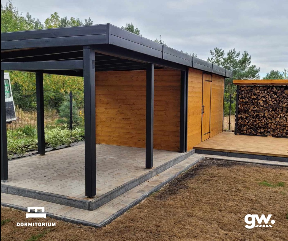 Domek narzędziowy z zabudową na drewno KVH i zadaszonym tarasem z kostki brukowej, widok z zewnątrz na tle drzew.