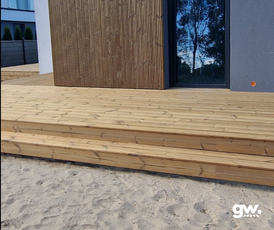 Wykonany taras z jasnego drewna z dwoma stopniami, przylegający do nowoczesnego domu z elementami elewacji z pionowych desek i dużym oknem z refleksami drzew.