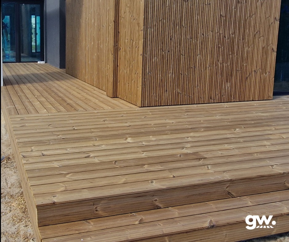Wykończony taras z desek thermososna z trzema stopniami, przylegający do budynku z elewacją z pionowych desek, widok z bliska na strukturę drewna.