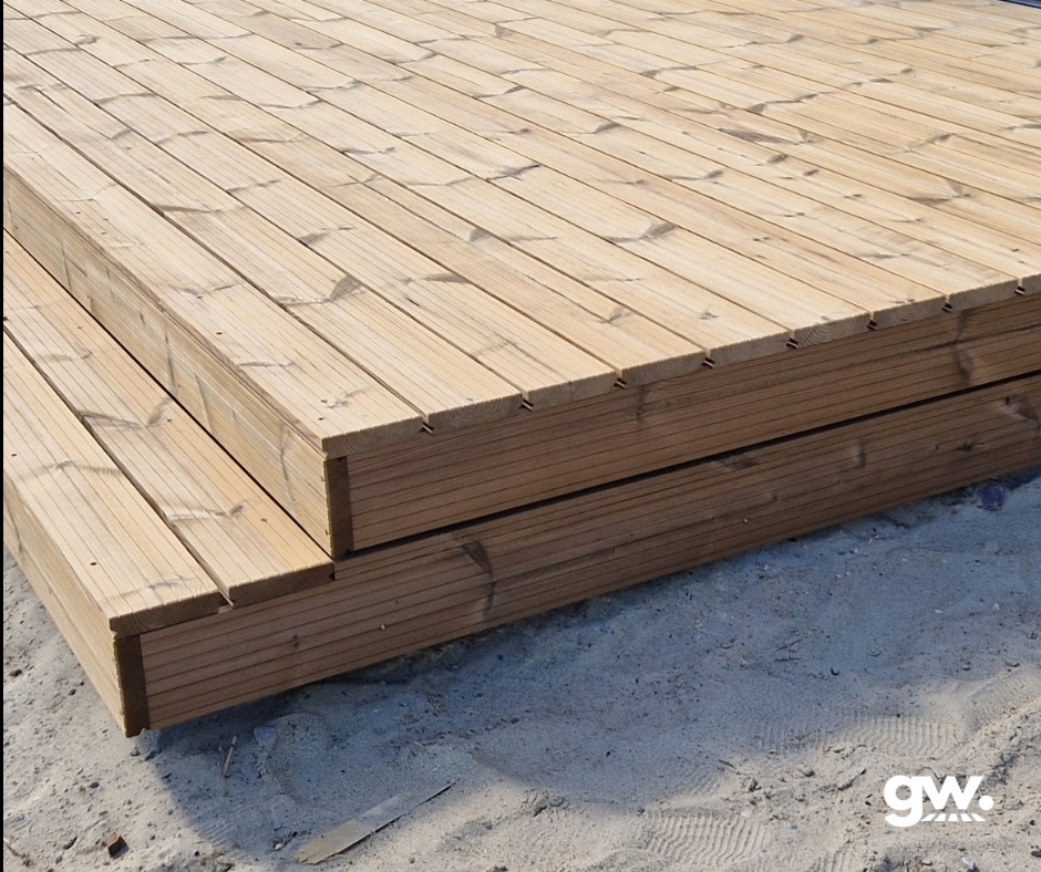 Nowo wybudowany taras z jasnego drewna thermososna, widoczne deski ułożone równolegle, zarys piasku w tle, logo w prawym dolnym rogu.