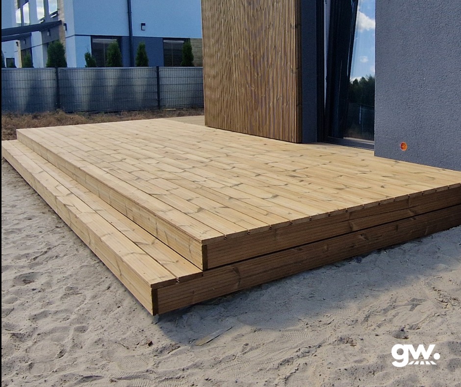 Świeżo wykonany taras z jasnego drewna, z dwoma stopniami, przylegający do budynku z elewacją z pionowych desek i szarego tynku, otoczony piaszczystym podłożem.