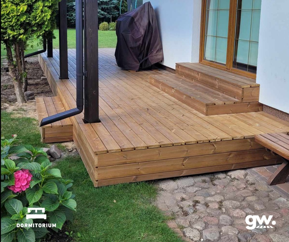 Wykonany taras z desek thermososna, z widocznymi schodkami prowadzącymi do drzwi, czarnymi rynnami i fragmentem ogrodu z hortensją.