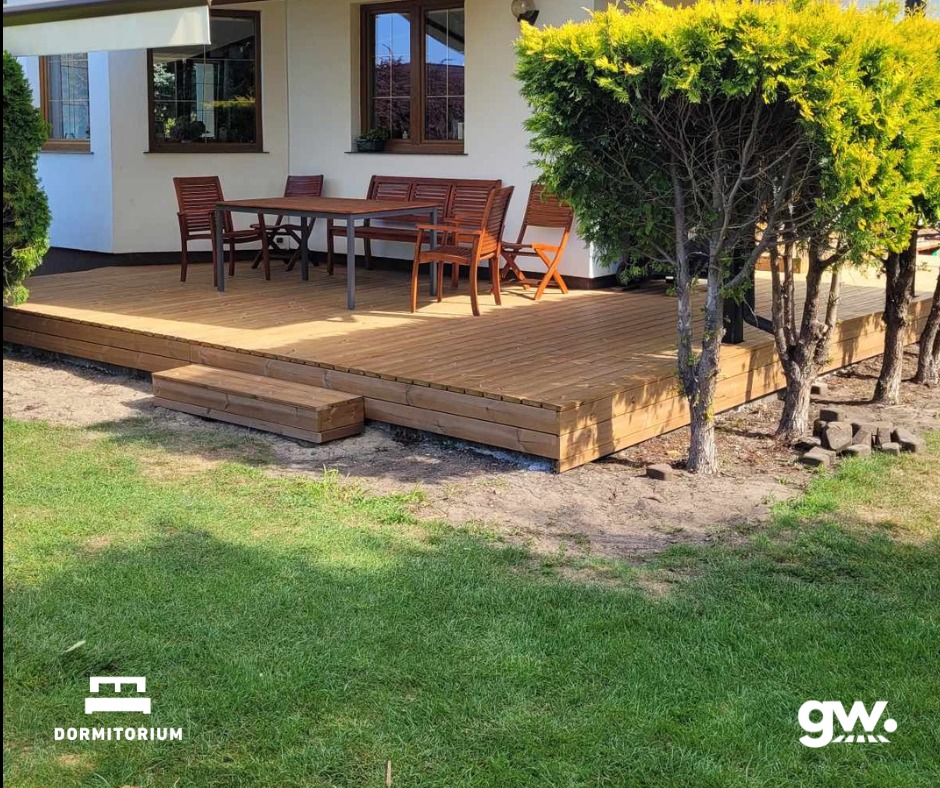 Jasny taras z drewna thermososna, z niskimi schodkami wejściowymi, zestawem mebli ogrodowych i drzewami tuż obok, na tle białego domu z brązowymi oknami. W dolnych rogach widoczne logotypy firmy.