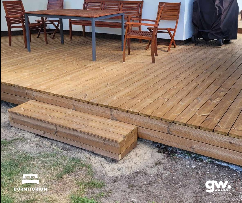 Jasny taras z drewna thermososna z niskimi schodami wejściowymi, zestaw mebli ogrodowych z drewna i metalu, grill pod pokrowcem w tle.