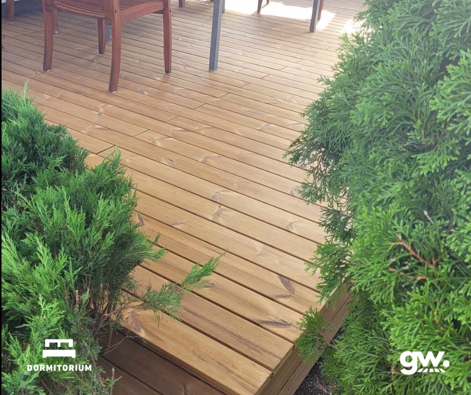 Starannie wykonany taras z desek thermososna, otoczony zielonymi krzewami iglastymi, z widocznym fragmentem mebli ogrodowych w tle.