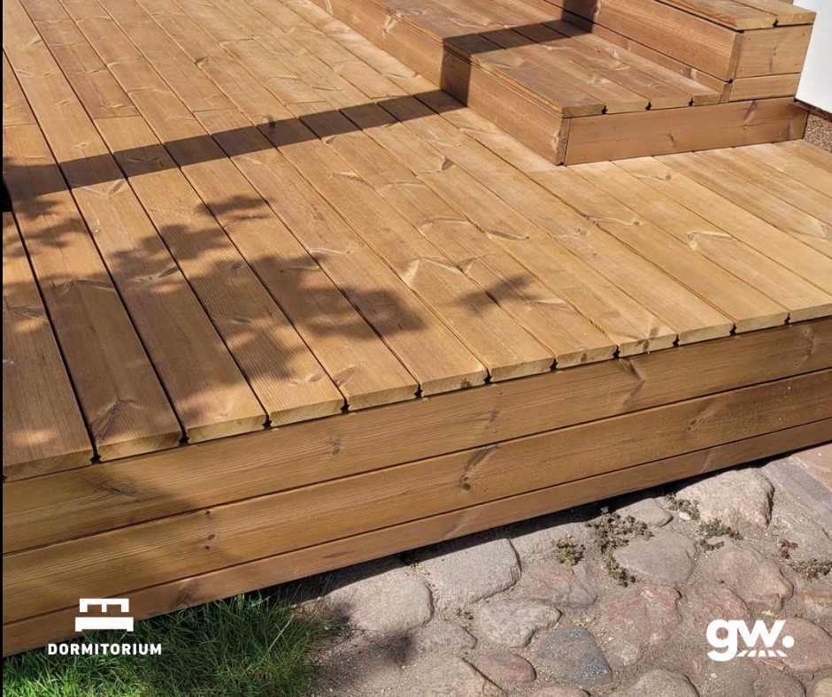 Drewniany taras z widocznymi schodkami, wykonany z desek thermososna, z cieniem drzewa i fragmentem kamiennego podłoża.