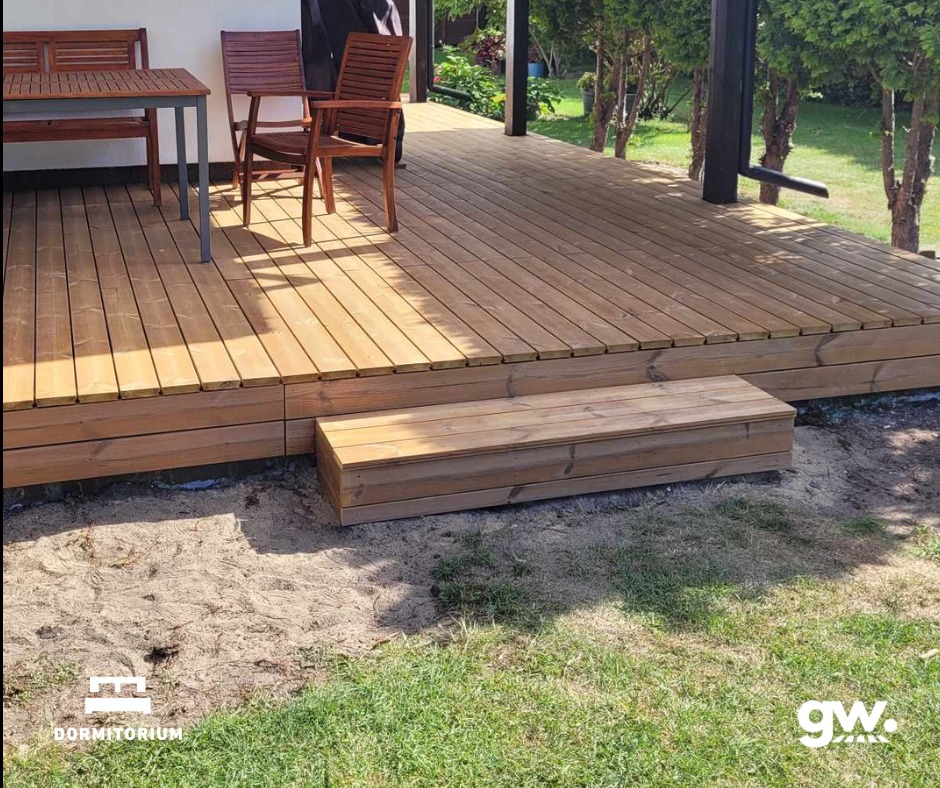 Wykończony taras z deski thermososna, z drewnianymi meblami ogrodowymi, widok na trawnik i zieleń w tle.