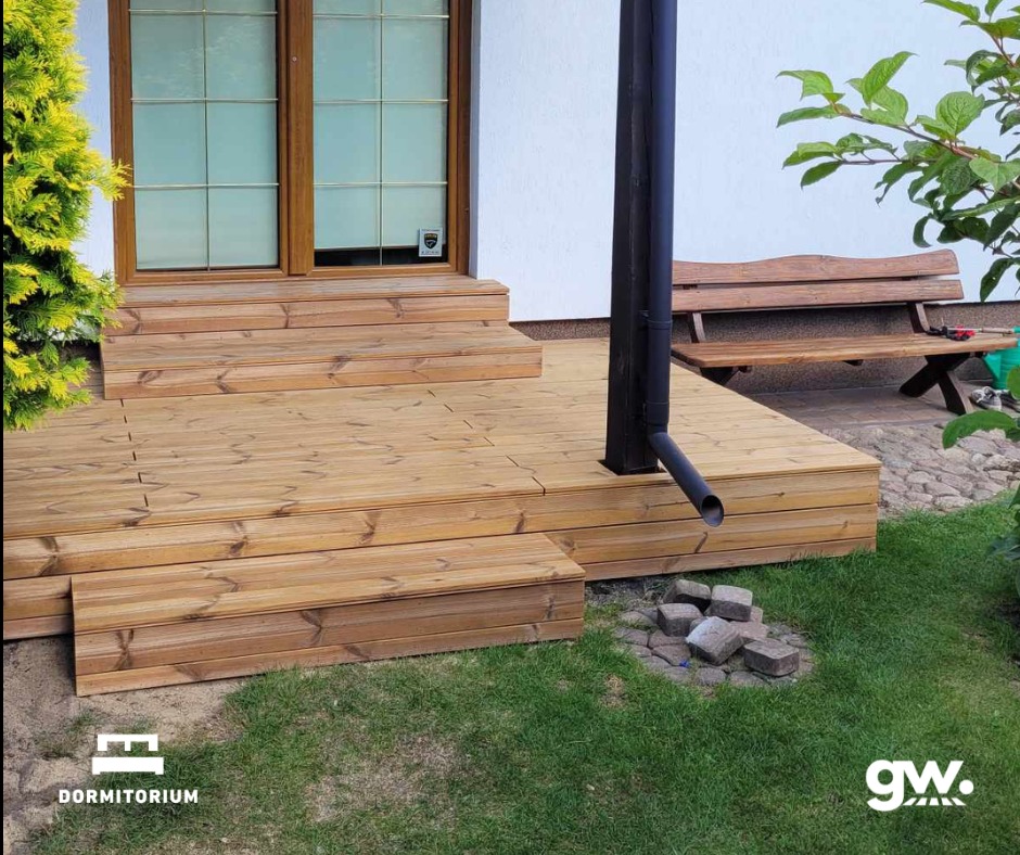 Nowo wybudowany taras z jasnego drewna z widocznymi schodkami, rynną odprowadzającą wodę i ławką w tle, otoczony zielenią.