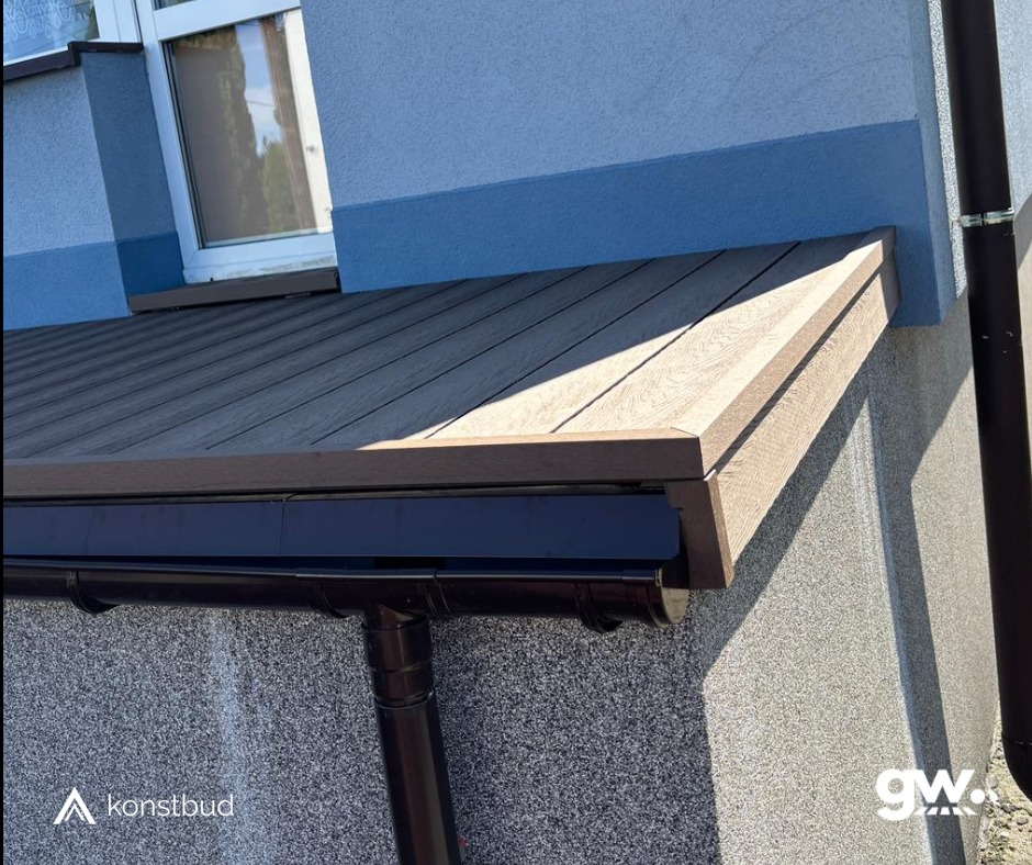 Fragment elewacji budynku z wykończonym zadaszeniem nad oknem, wykonanym z desek kompozytowych w odcieniach brązu, z widoczną rynną i rurą spustową.