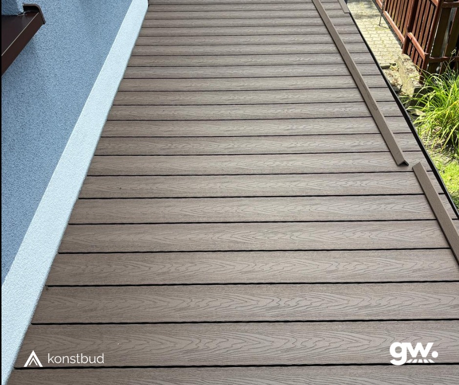Ułożone deski tarasowe z kompozytu w odcieniach brązu, widoczne logo firmy na dole zdjęcia, listwa wykończeniowa przy ścianie budynku.