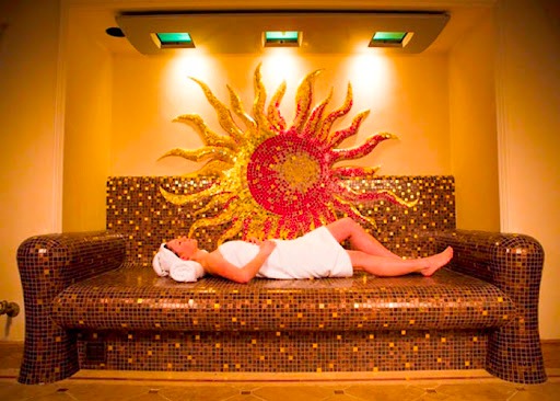 Mozaikowa leżanka w SPA z dekoracją ścienną w kształcie słońca, podświetlona lampami, na której leży kobieta owinięta ręcznikiem.