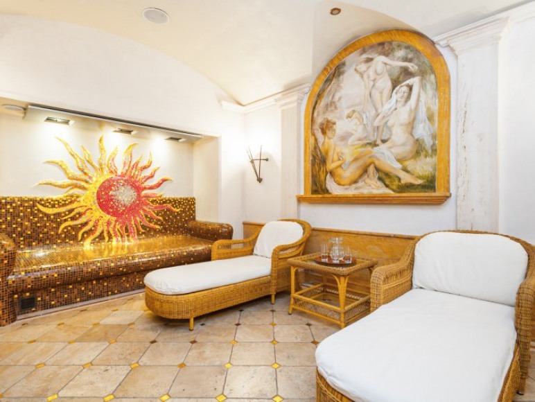 Relaksacyjna strefa SPA z mozaikową sofą w kształcie słońca, wiklinowymi leżankami i klasycznym malowidłem na ścianie w łukowej niszy.