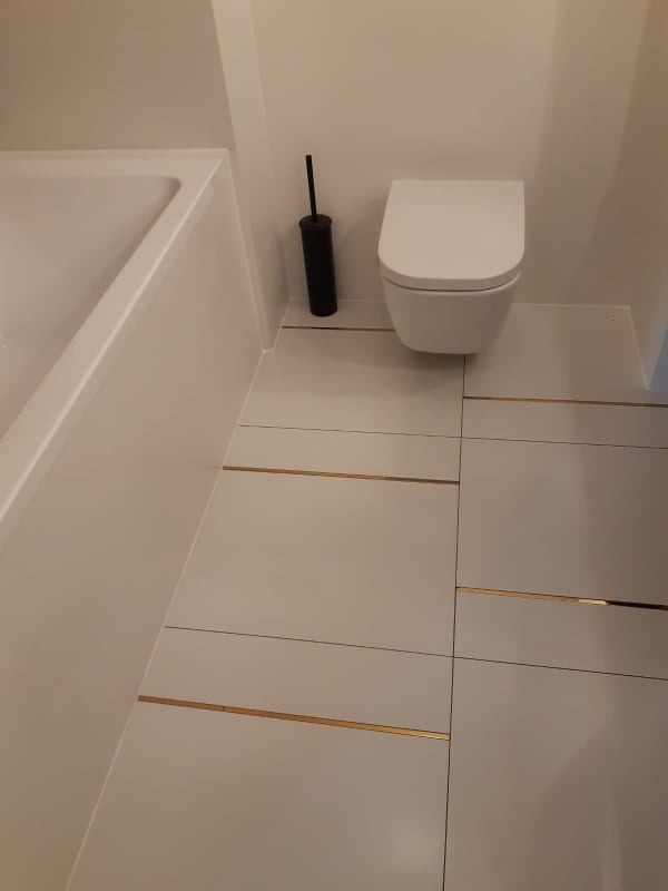 Nowoczesna łazienka z białymi płytkami podłogowymi z subtelnymi złotymi wstawkami, wisząca toaleta i czarny szczotka do WC.