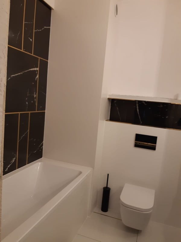 Nowoczesna łazienka w bieli i czerni z akcentami złota: wanna z czarnymi płytkami imitującymi marmur, wisząca toaleta i akcesoria łazienkowe, minimalistyczny design.