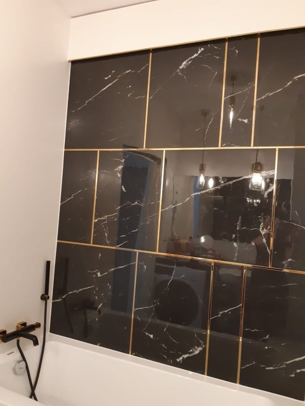 Część łazienki z wanną i czarnymi płytkami imitującymi marmur, przedzielonymi złotymi listwami, widoczna czarna bateria wannowa