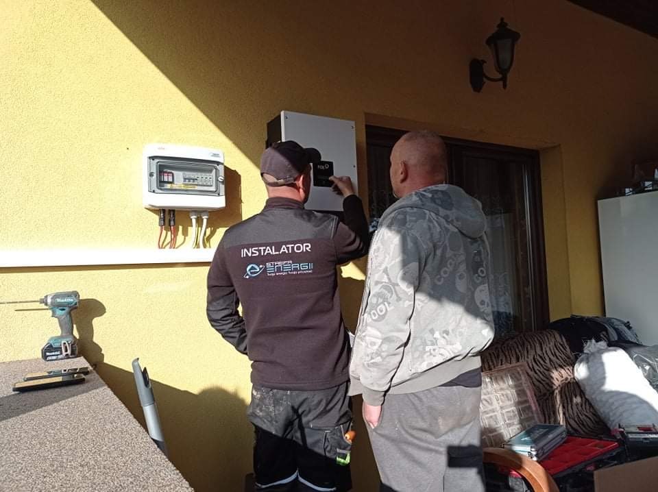 Dwóch mężczyzn instaluje białą skrzynkę i licznik na żółtej ścianie budynku; jeden z nich w czarnej bluzie z napisem 'Instalator Strefa Energii'.