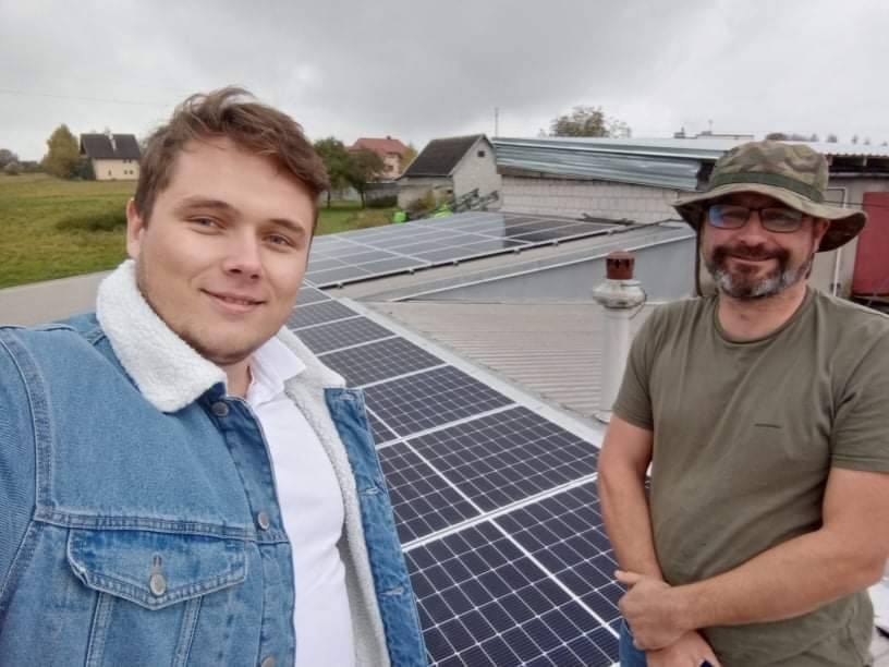 Dwaj mężczyźni, jeden w dżinsowej kurtce z kożuszkiem, drugi w kapeluszu i okularach, stoją na dachu z panelami słonecznymi w pochmurny dzień.