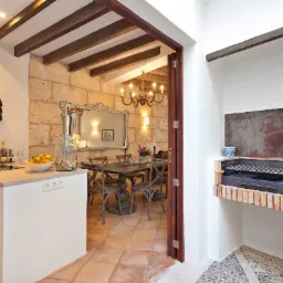 Wnętrze kuchni przechodzącej w jadalnię z grillem wbudowanym w ścianę, kamienne ściany, drewniane belki sufitowe, rustykalne meble, dekoracyjna półka w kształcie koguta.