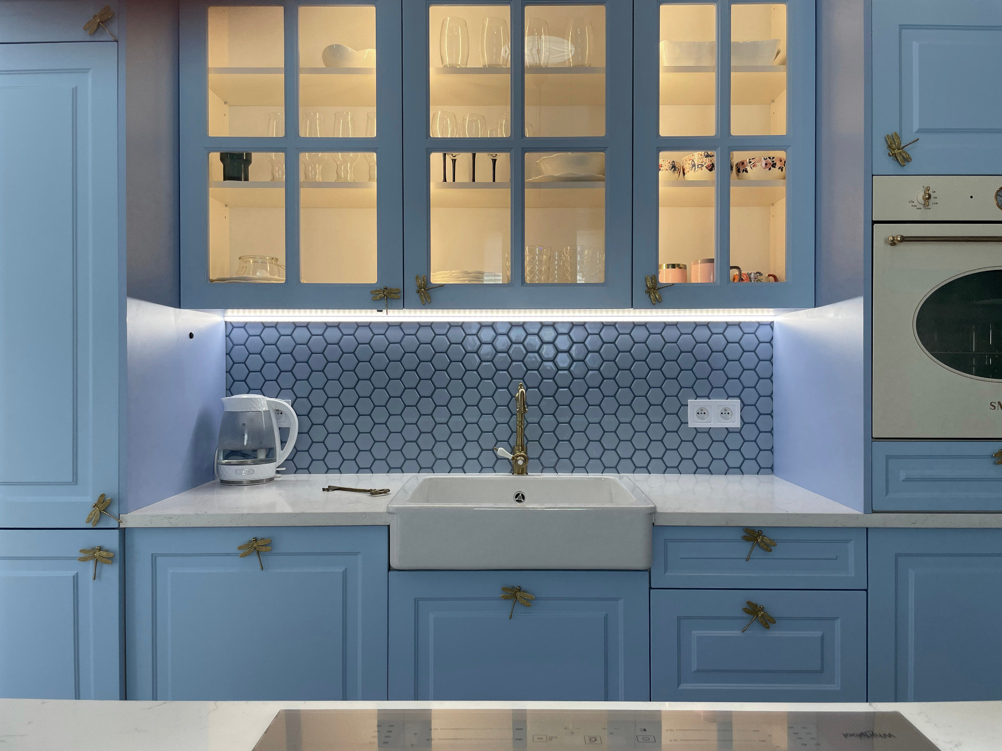 Błękitna kuchnia w stylu rustykalnym: szafki z przeszkleniami, armatura w kolorze złota, heksagonalne płytki, uchwyty w kształcie ważek, czajnik elektryczny.