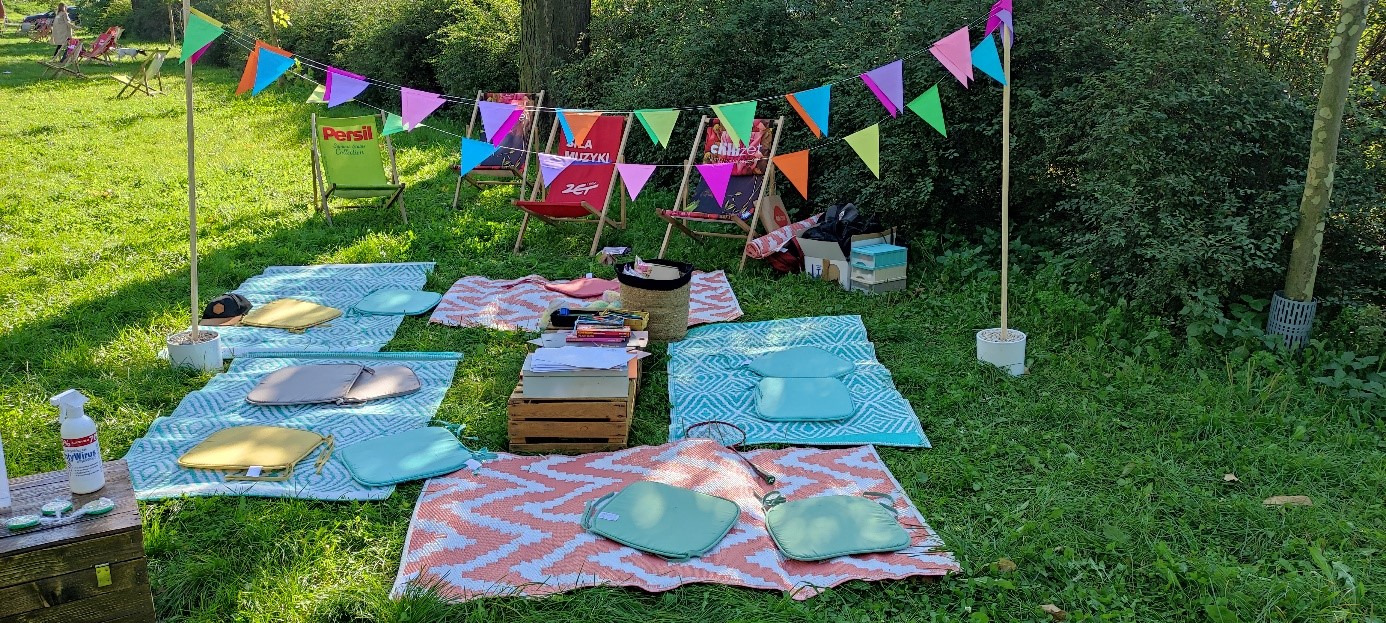 Uroczystość plenerowa z kolorowymi proporczykami, rozłożonymi kocami, poduszkami, leżakami i stolikiem z książkami, przygotowana na trawie w otoczeniu zieleni.