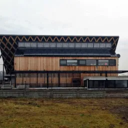 Nowoczesny dom z panelami fotowoltaicznymi na dachu, drewnianą elewacją i nietypową konstrukcją dachu w kształcie piły, widok z zewnątrz w pochmurny dzień.