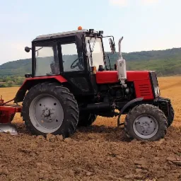 Czerwony traktor podczas orki pola, z widocznym pługiem i świeżo przekopaną ziemią, tło stanowią pola i zalesione wzgórza.