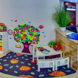 Kolorowy kącik zabaw dla dzieci w poczekalni z naklejkami sowy i drzewa na ścianie, stolik z książeczkami i dywan z motywem sów.