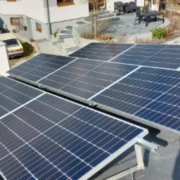 Widok z góry na panele fotowoltaiczne zamontowane na dachu domu, z widocznym ogrodem i elementami architektury w tle.