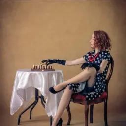 Kobieta w sukience w grochy i czarnych rękawiczkach siedzi przy stoliku z szachami, stylizowana sesja.