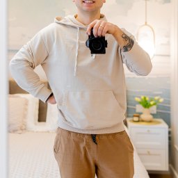 Krycha Fotografuje - Uśmiechnięty mężczyzna w beżowej bluzie z kapturem, robiący sobie zdjęcie w lustrze, w tle widoczny pokój z łóżkiem i kwiatami.