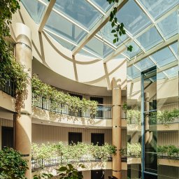 Krycha Fotografuje - Wnętrze hotelu z przeszklonym dachem, kolumnami i balkonami porośniętymi zielenią. Widoczna winda panoramiczna i światło słoneczne wpadające przez dach.