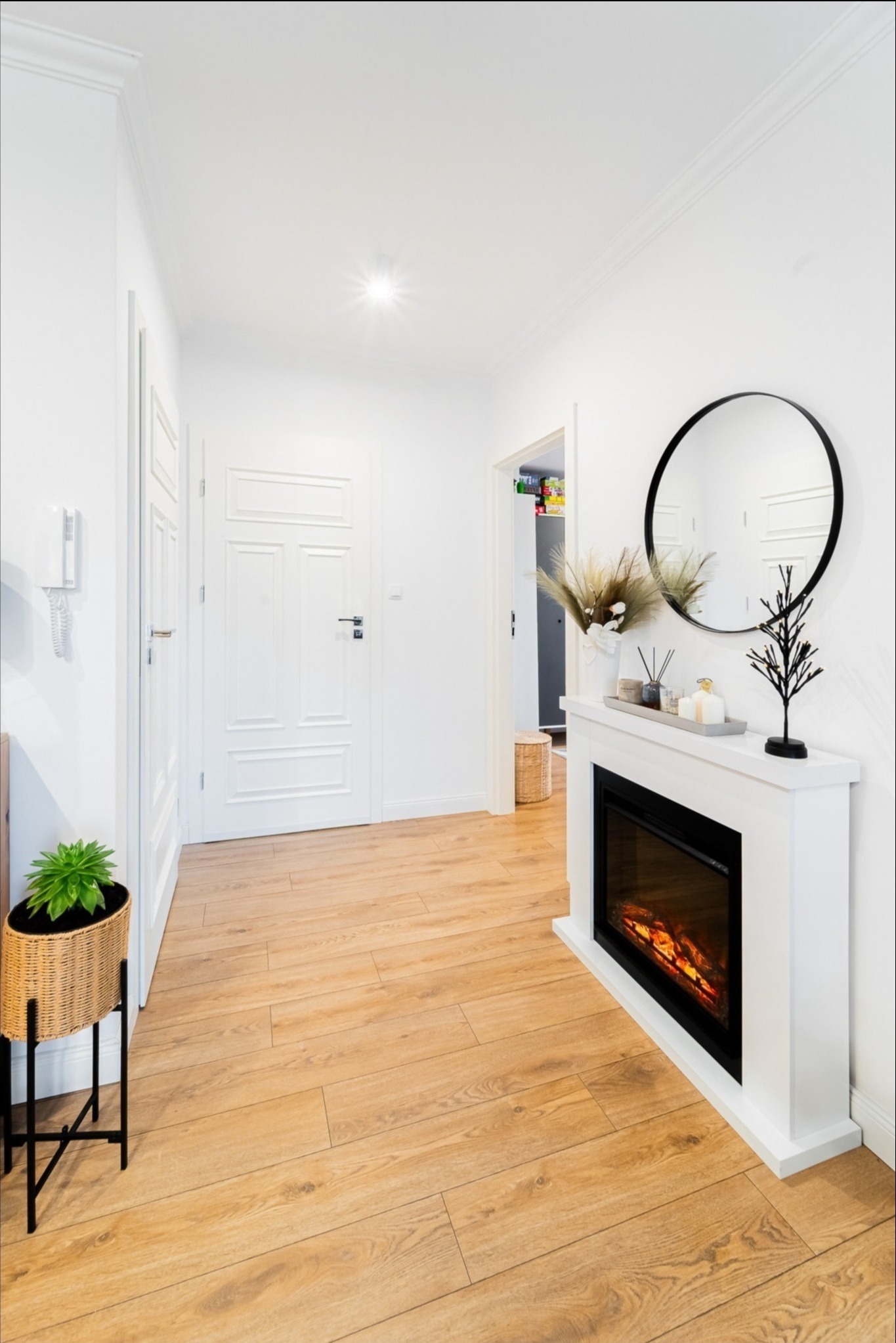 Jasny korytarz z drewnianą podłogą, białymi drzwiami i kominkiem elektrycznym zwieńczonym okrągłym lustrem. W rogu stoi pleciony kwietnik z zieloną rośliną.