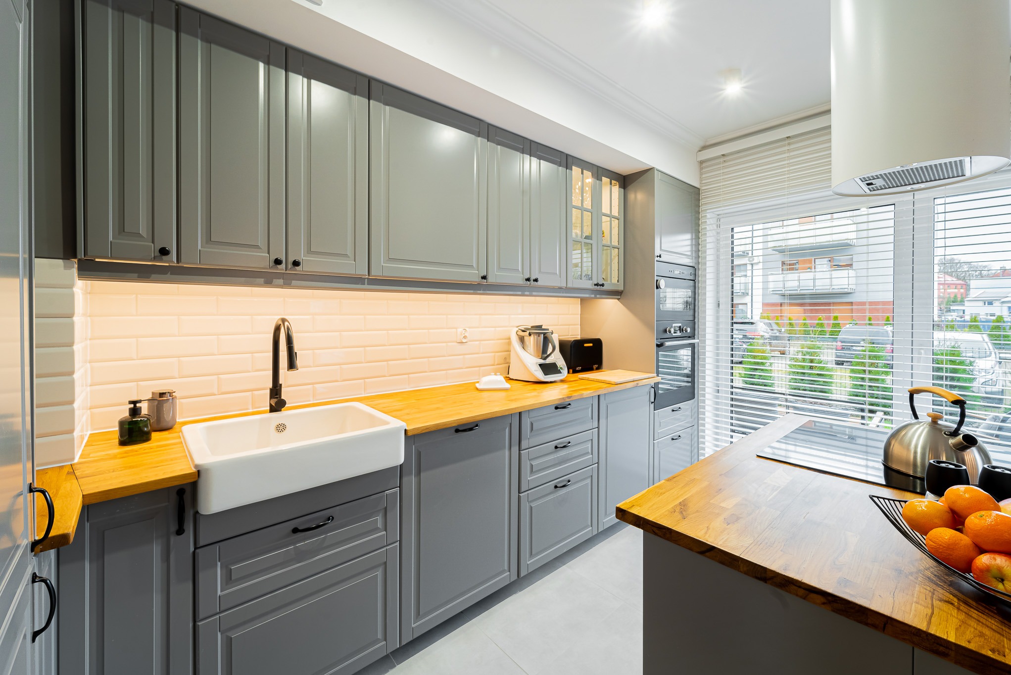 Szara kuchnia z drewnianym blatem, białym zlewem farmerskim i ceglanymi płytkami. Nowoczesny design z widokiem na zewnątrz przez żaluzje.